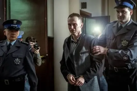 Scena z filmu o Tomaszu Komendzie: policjanci eskortują go w kajdankach, a za nimi stoi fotograf z aparatem.