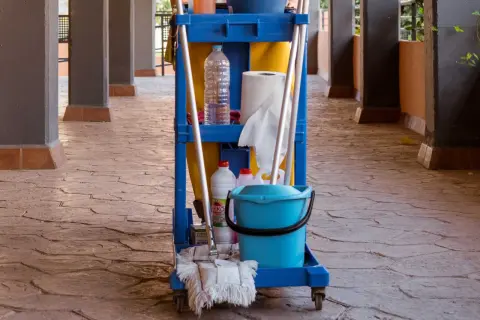 Carro de limpieza con cubeta azul, botellas y escoba sobre un suelo poroso de piedra.