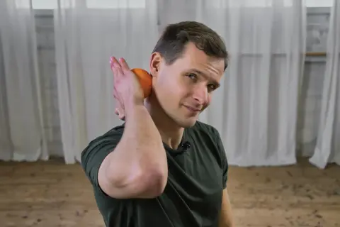 Mann lockert Nackenmuskulatur mit einer Orange.