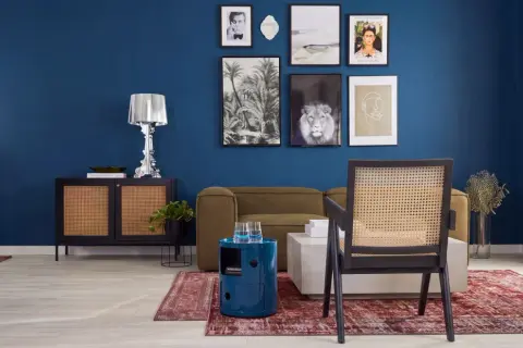 Ein stilvolles Wohnzimmer mit einer tiefblauen Wand, die mit Kunstwerken geschmückt ist. Ein Rattan-Stuhl steht vor einem Sofa, daneben ein runder, blauer Beistelltisch.