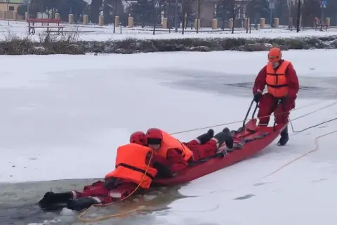 Ratownicy w akcji, wyciągają osobę z lodowatej wody za pomocą **sanki ratownicze**. Zimowa sceneria i profesjonalny sprzęt.