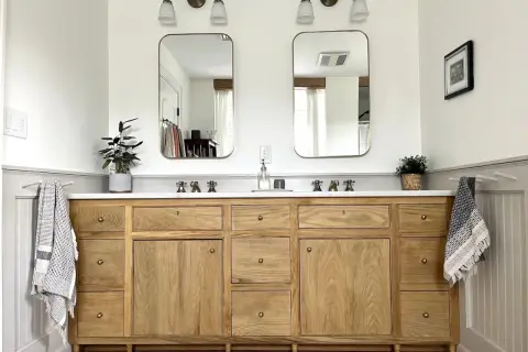 A beautiful DIY bathroom vanity with a natural wood finish, marble countertop, and brass fixtures. Two mirrors hang above, reflecting the bright, airy space.