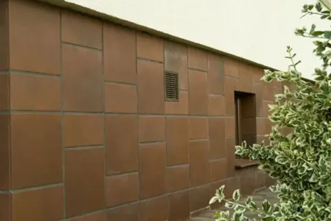 Brązowy cokół budynku, wyłożony płytkami o nieregularnym układzie, z małym okienkiem i kratką wentylacyjną.