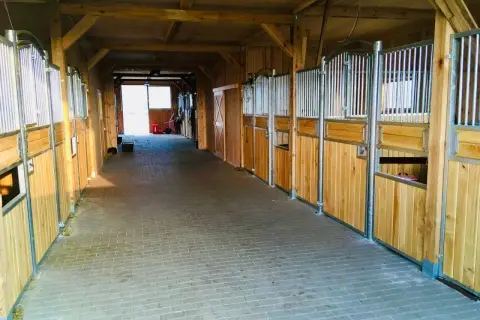 Wnętrze stajni w stadninie koni Dolina Pilicy. Drewniane boksy dla koni, wyłożona kostką brukową alejka, światło wpada przez otwarte drzwi na końcu.