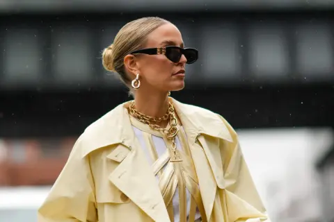 Kobieta w okularach przeciwsłonecznych i żółtym płaszczu, z eleganckim upięciem włosów w kok. Złota biżuteria dodaje szyku.