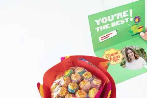 En person håller upp ett kort med texten "YOU'RE THE BEST" och en bild på sig själv. Bredvid syns en bukett med Chupa Chups-klubbor, en fin godis present diy.
