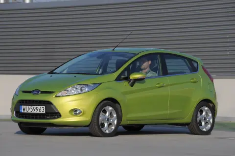 Zielony Ford Fiesta Mk7 z widocznymi żarówkami z przodu, zaparkowany przed budynkiem.