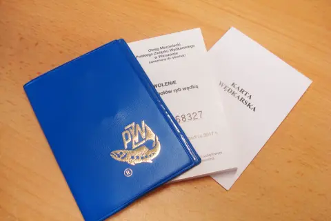 Niebieska książeczka z logo PZW i dokumenty "Karta Wędkarska" na drewnianym tle.