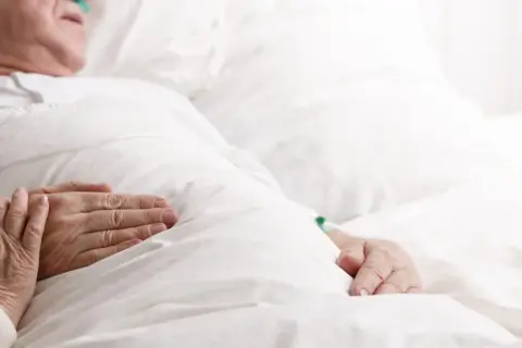 Dłonie starszej osoby spoczywają na białej pościeli, czuć delikatny oddech biota.