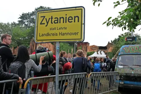 Besucher stehen vor einem Schild "Zytanien statt Lehrte, Landkreis Hannover". Ein bunt bemalter Van und Festivalbauten sind im Hintergrund zu sehen.