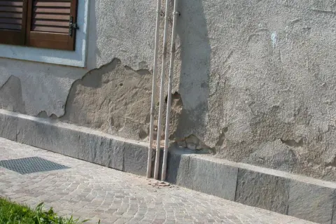 Muro esterno che si sbriciola, mostrando crepe e intonaco scrostato vicino a tubi verticali e una finestra con persiane.