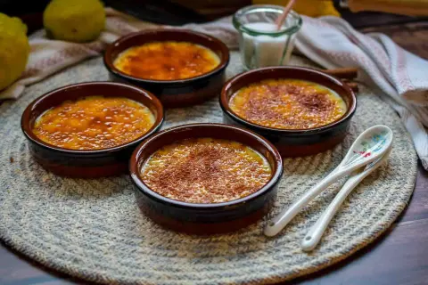 Vier Crema Catalana Schalen mit karamellisierter Zuckerkruste, bereit für das Crema Catalana Rezept. Zitronen und Löffel liegen daneben.