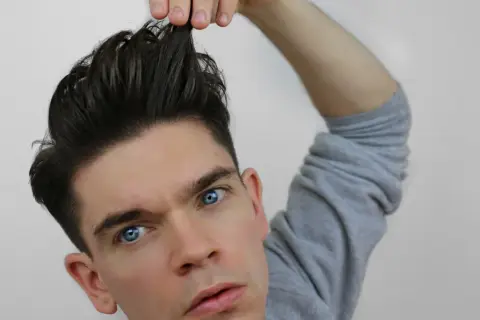 Mann mit einem messy quiff, der sich die Haare hochzieht. Blaue Augen, graues Sweatshirt.