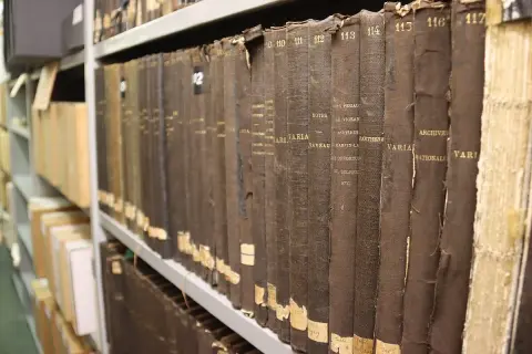 Rangées de vieux registres reliés en tissu brun dans les archives départementales 85. Les titres sont partiellement lisibles.