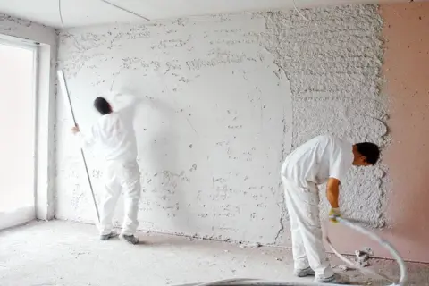 Operai in tuta bianca applicano intonaco grezzo alle pareti, preparando la finitura a civile.