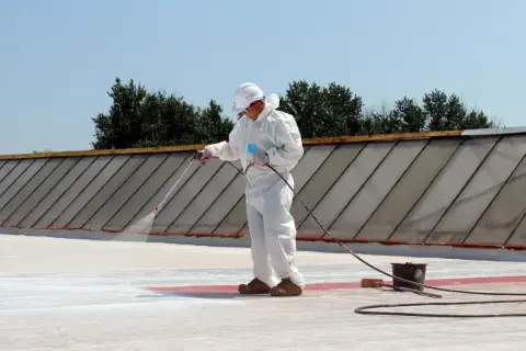 Pracownik w białym kombinezonie natryskuje nowoczesne materiały na dach płaski EPDM, porównując je z papą termozgrzewalną.