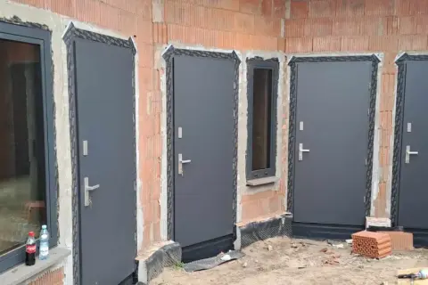 Nowe, szare drzwi zewnętrzne i okno w budowie. Widać ceglane ściany i materiały budowlane.