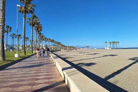 Paseo marítimo con palmeras en el **parque Roquetas de Mar**. Gente camina por la senda adoquinada junto a la playa de arena bajo un cielo azul.