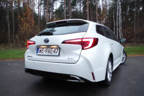 Biała Toyota Corolla 1.8 Hybrid na tle jesiennego lasu. Widok z tyłu, z podświetlonymi światłami.