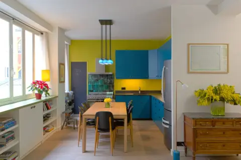 Plan cuisine ouverte sur salle à manger. Meubles bleus vifs, murs jaune citron, table en bois, chaises noires.
