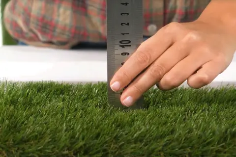 Mano mide la altura del césped artificial con una regla metálica. El césped es verde y denso.