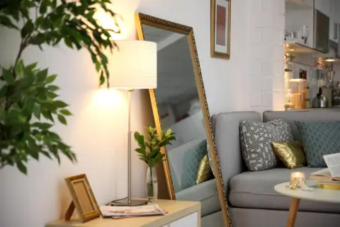 Ein großer Spiegel und eine Stehlampe machen die dunkle Wohnung heller. Ein Sofa mit Kissen und ein kleiner Tisch mit Kerze sind zu sehen.