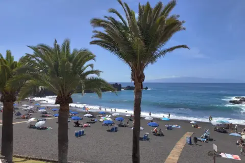 Palmy nad czarną plażą w Puerto de Santiago, gdzie turyści odpoczywają pod parasolami.
