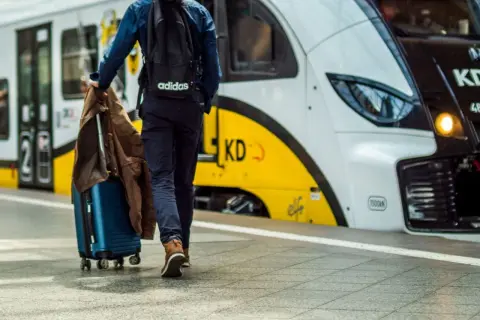 Podróżny z walizką i kurtką idzie w stronę pociągu KD. Rozkład PKP czeka na pasażerów.