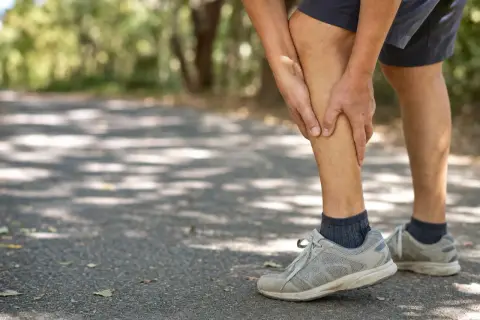 Uomo con dolore alle gambe, difficoltà a camminare su un sentiero ombreggiato.