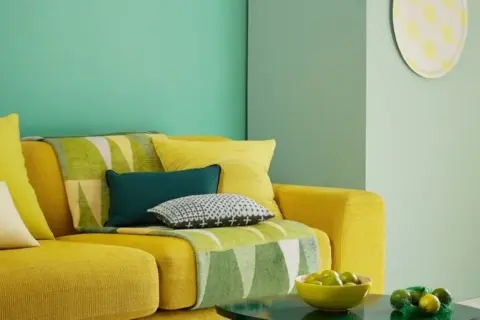 Divano giallo acceso con cuscini colorati e una coperta con motivi geometrici in **colore verde**. Sul tavolino, una ciotola di lime.