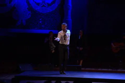 Un bailaor flamenco español actual, con camisa blanca y pañuelo, baila en el escenario. A su lado, dos cantantes y un guitarrista.
