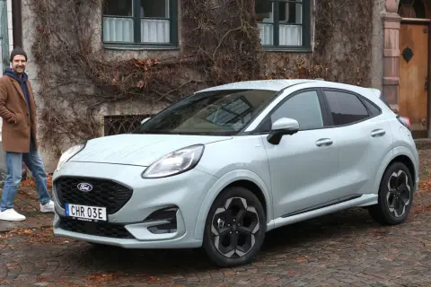 Man står bredvid Fords minsta modell, en ljusgrå Puma, framför ett gammalt hus med murgröna.