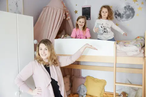 Uroczy mały pokój dla rodzeństwa w bloku. Dwie dziewczynki bawią się na łóżku piętrowym, a mama prezentuje przestrzeń.
