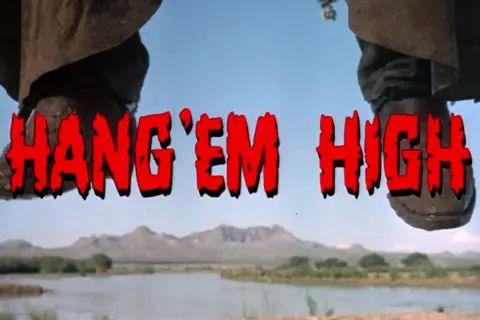 Nogi w kowbojkach wiszą nad rzeką. Czerwony napis "HANG 'EM HIGH" sugeruje westernowe porachunki.