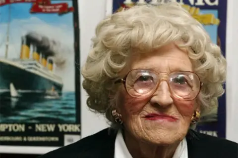 Una anciana con gafas grandes sonríe. Detrás de ella, carteles del Titanic, evocando a los supervivientes del trágico viaje.