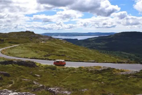 Eine malerische England Landschaft mit einer kurvigen Straße, einem roten Campervan und einem See in der Ferne unter bewölktem Himmel.