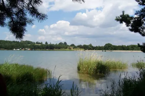 Spokojny zalew Dźibice otoczony zielenią, z kempingiem na brzegu i chmurami na niebie.