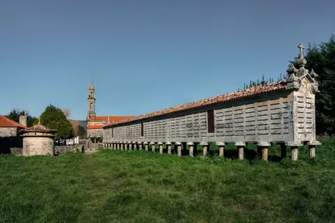 El horreo de Carnota, una estructura de piedra sobre pilares, se alza majestuoso ante una iglesia y un cielo azul.