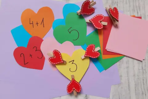 Kolorowe serca z cyframi i działaniami matematycznymi, ozdobione drewnianymi klamerkami w kształcie serc.