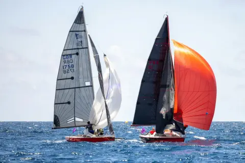 Due barche a vela competono in mare aperto. La barca a sinistra ha una vela grigia con numeri e simboli, mentre quella a destra sfoggia vele nere e arancioni. Lo strallo, elemento cruciale per la stabilità, è ben visibile.