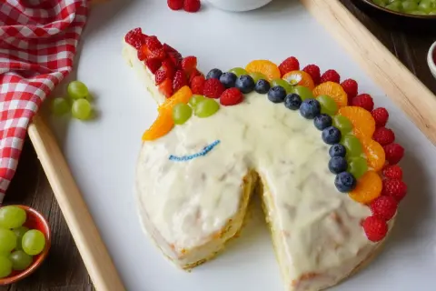 Ein fantasievoller Kuchen ohne Zucker in Einhornform, verziert mit bunten Früchten wie Erdbeeren, Blaubeeren und Mandarinen.