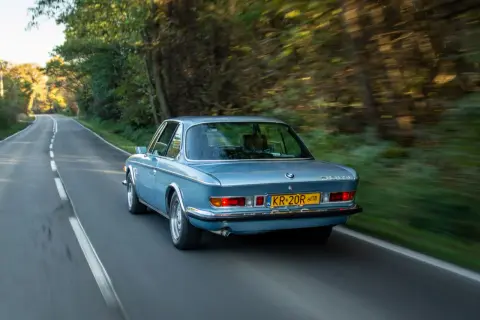 Niebieskie BMW E9 z lat 70. sunie po leśnej drodze, tworząc wrażenie prędkości.