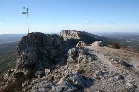 Vue panoramique depuis le pic des mouches, avec un anémomètre sur un rocher et un paysage de montagnes et de vallées sous un ciel bleu.