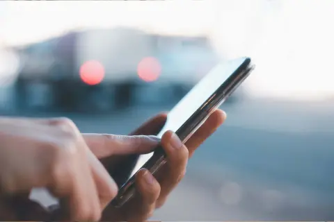 Dłoń dotyka ekranu smartfona, szukając informacji, jak przywrócić ustawienia fabryczne w telefonie. W tle rozmyte światła.
