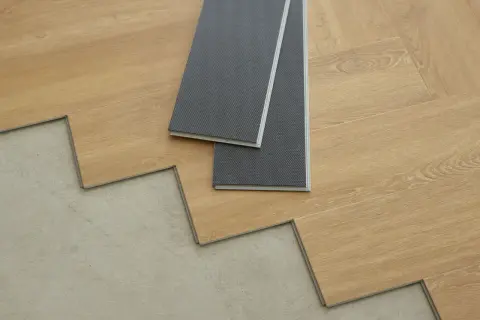 Panele winylowe w trakcie układania na betonowej podłodze. Dwa szare panele leżą na drewnopodobnych deskach.