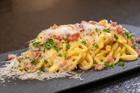 Eine Portion Spaghetti Carbonara mit Speckwürfeln, Petersilie und Parmesan auf einem dunklen Teller.