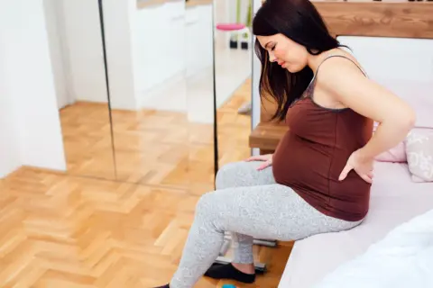 Kobieta w 27 tygodniu ciąży siedzi na łóżku, masując bolące plecy.