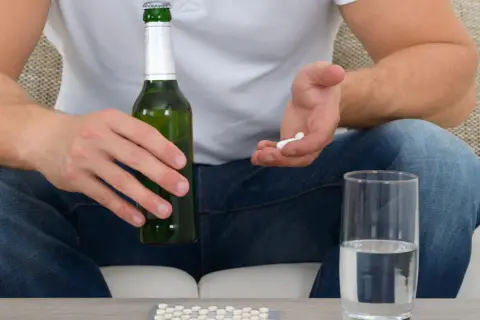 Mężczyzna trzyma butelkę piwa i garść tabletek, obok stoi szklanka wody. Czy to ibuprofen i alkohol?