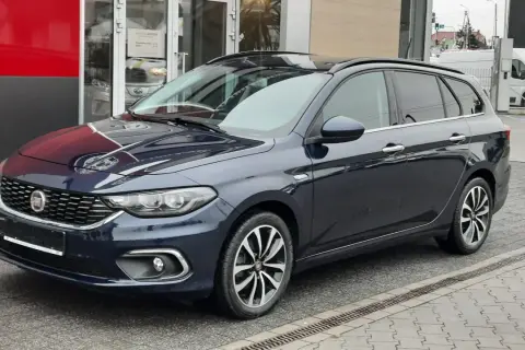 Ciemnoniebieskie kombi Fiat Tipo stoi przed budynkiem z czerwonym i szarym panelem.
