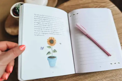 Een open dagboek met een zonnebloem en vlinder illustratie. Een roze pen ligt op de rechterpagina, klaar om gedachten uit je hoofd op te schrijven.